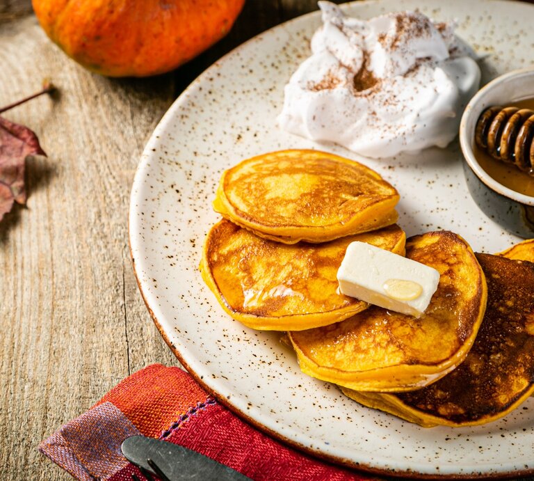 Vier Kürbis Pancakes auf einem hellen Teller mit Honig und etwas geschlagener Sahne. Alles von schräg oben auf einem rustikalen Holztisch mit herbstlicher Deko fotografiert. | © Gettyimages/ a_namenko 