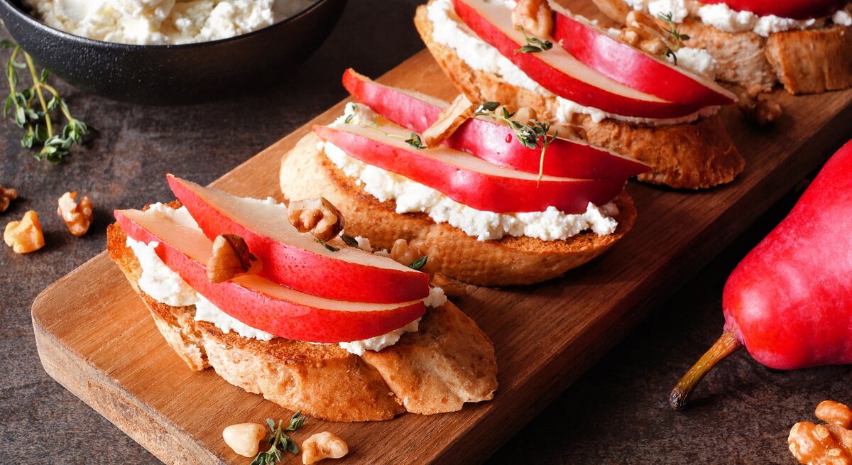 Draufsicht schräg: Ein Holzbrettchen mit vier Hüttenkäse-Sandwich mit Birne und Walnüssen liegt auf einem dunklen Holzuntergrund. Im Hintergrund ist eine dunkle Schüssel mit Hüttenkäse. Neben dem Brettchen ist ein Teil einer roten Birne zu sehen.