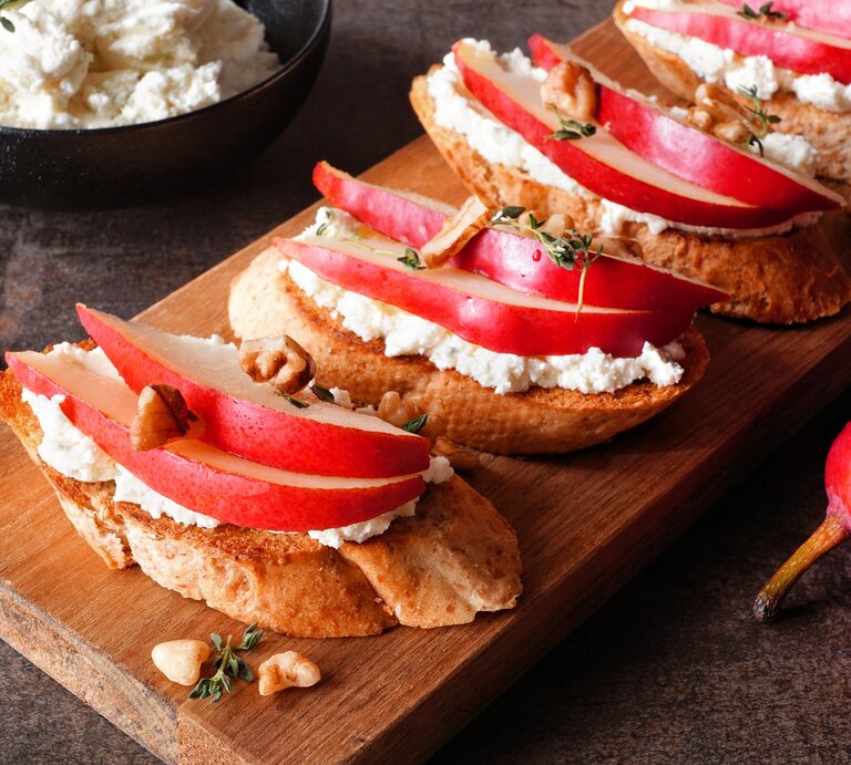 Draufsicht schräg: Ein Holzbrettchen mit vier Hüttenkäse-Sandwich mit Birne und Walnüssen liegt auf einem dunklen Holzuntergrund. Im Hintergrund ist eine dunkle Schüssel mit Hüttenkäse. Neben dem Brettchen ist ein Teil einer roten Birne zu sehen. | © Adobe Stock / Jenifoto