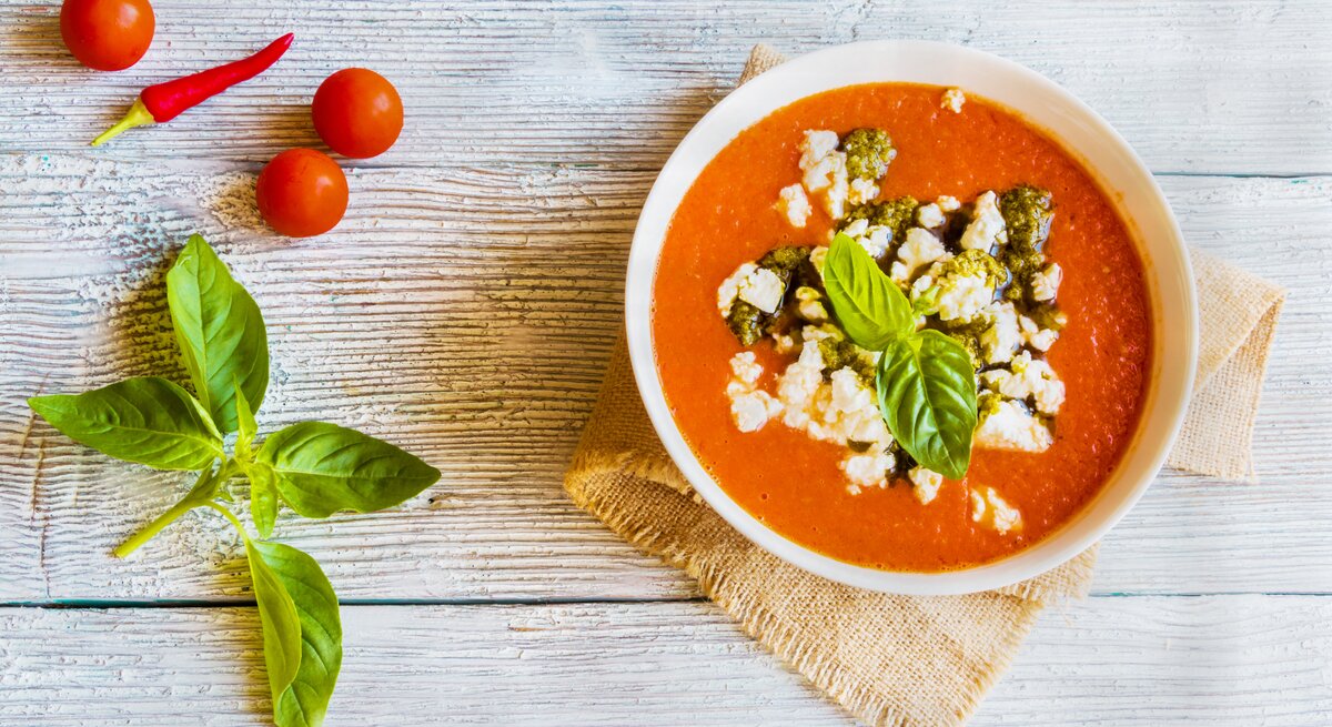 Rote Tomaten-Feta-Suppe mit Pesto und Basilikum in weüßer Schüssel auf Holz.