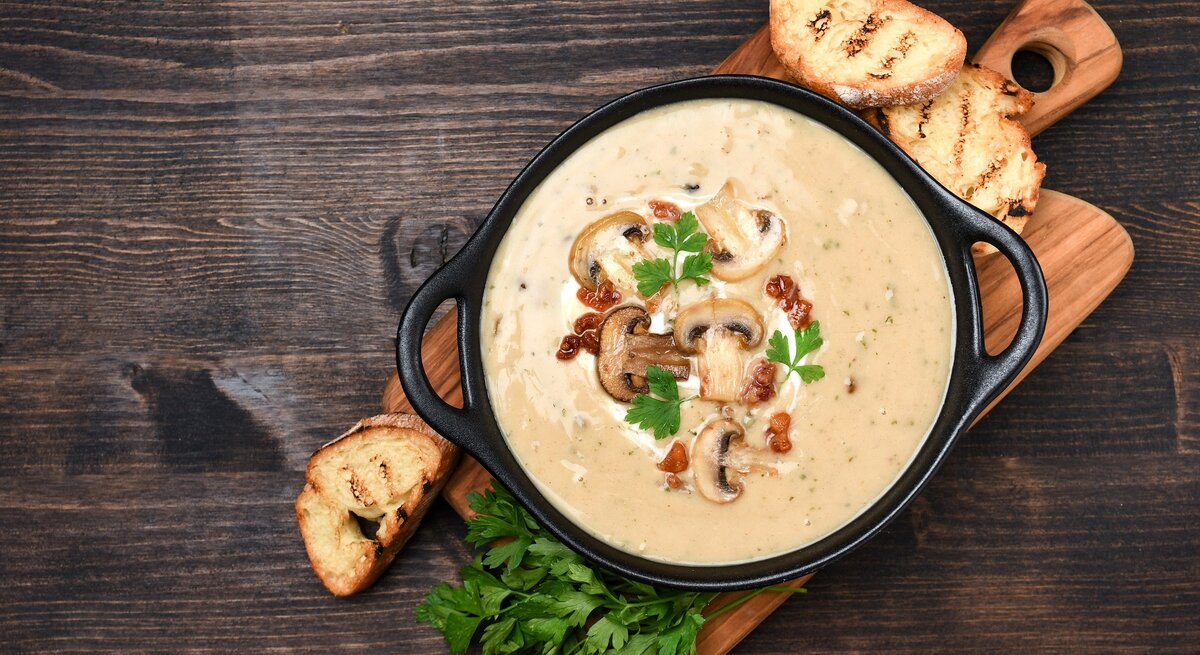 Draufsicht: Ein Holzbrettchen auf dem einen schwarze Schüssel steht. In der Schale ist eine alpenländische Kümmelrahmsuppe mit Pilzen. Drum herum liegen drei Scheiben geröstetes Weißbrot und frische Petersilie.