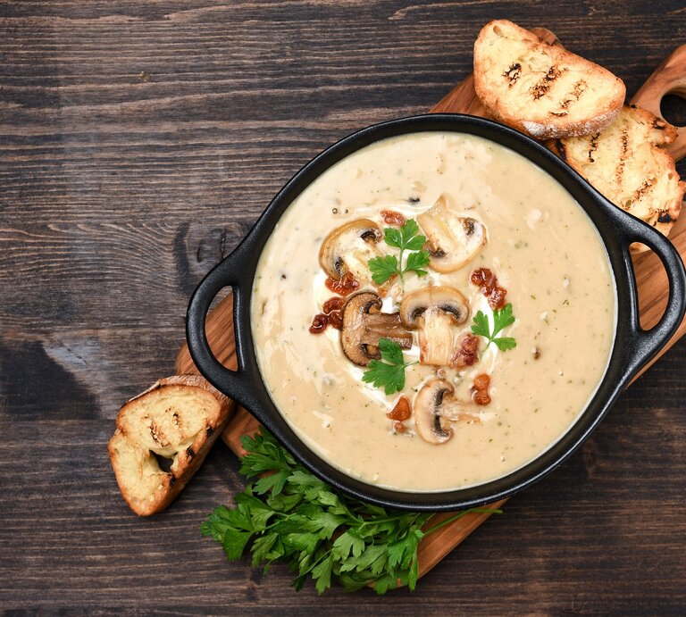 Draufsicht: Ein Holzbrettchen auf dem einen schwarze Schüssel steht. In der Schale ist eine alpenländische Kümmelrahmsuppe mit Pilzen. Drum herum liegen drei Scheiben geröstetes Weißbrot und frische Petersilie. | © Shutterstock / Elena.Katkova