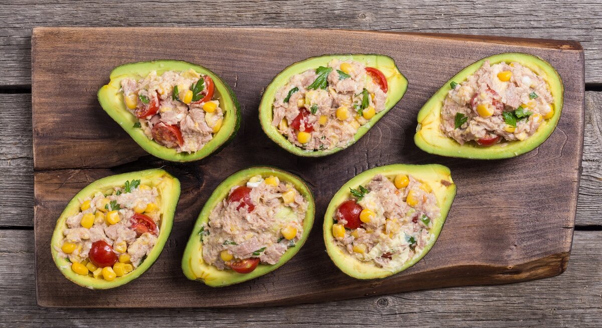 Ein Holzbrett mit sechs Avocadohälften und dem Thunfischsalat. Alles auf einem rustikalem Holztisch direkt von oben fotografiert.