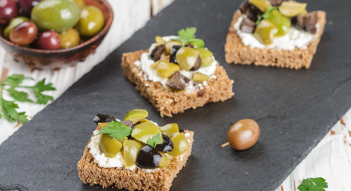 Draufsicht schräg: drei kleine Oliven-Feta-Stullen die auf einem Schiefer-Brettchen liegen. Daneben ist eine kleine Schale mit frischen Oliven.