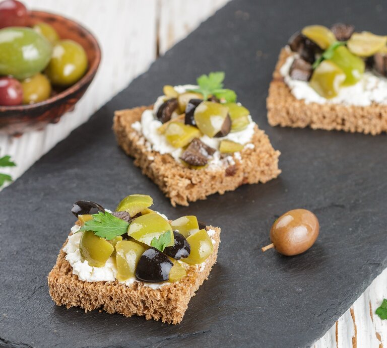 Draufsicht schräg: drei kleine Oliven-Feta-Stullen die auf einem Schiefer-Brettchen liegen. Daneben ist eine kleine Schale mit frischen Oliven. | © Shutterstock / Viktory Panchenko