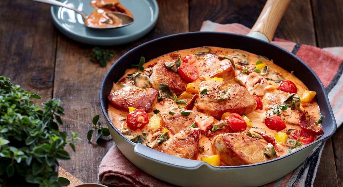 Puten-Tomaten-Pfanne in einer Pfanne auf Küchentuch auf Holzuntergrund. Drum herum Geschirr, ein Schälchen Salz und Kräuter. 