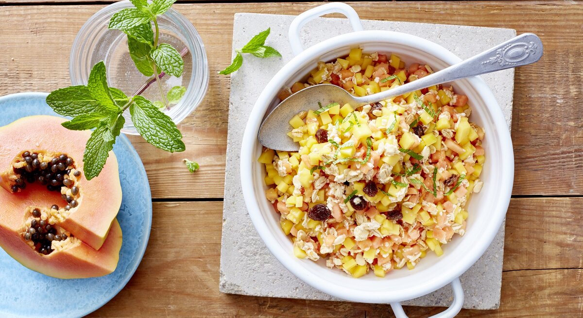Exotisches Müsli in einer weißen Schüssel auf einem hellgrauen Stein. Liegt neben frischer Minze und Papaya auf einem Holztisch