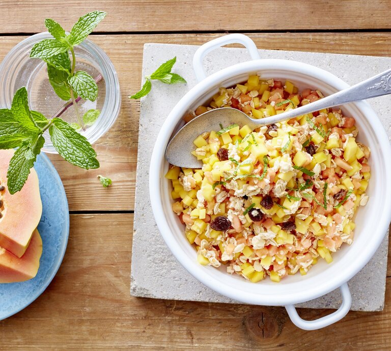 Exotisches Müsli in einer weißen Schüssel auf einem hellgrauen Stein. Liegt neben frischer Minze und Papaya auf einem Holztisch | © EAT CLUB/Isabell Triemer