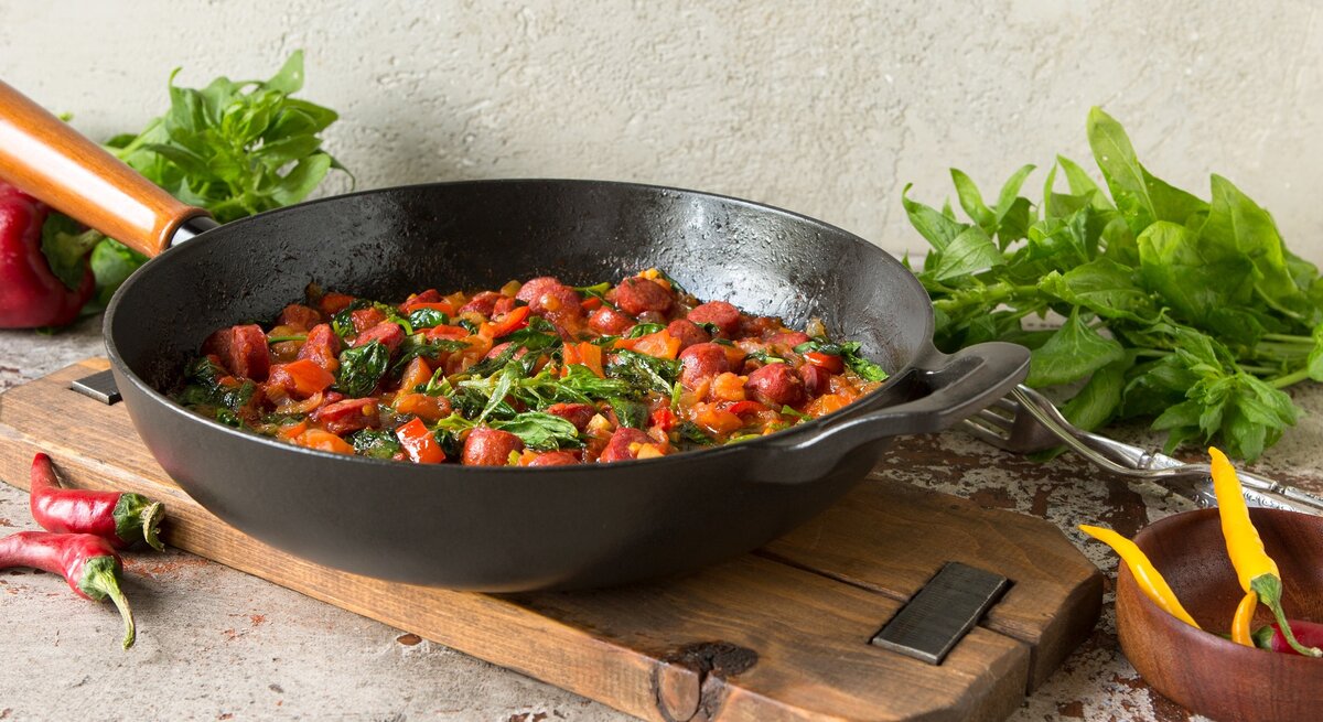 Frontal: Eine Würstchenpfanne mit Kidneys liegt auf einem rustikalen Holzbrett auf einem groben Untergrund. Drum herum liegen Chilis sowie frischer Spinat.