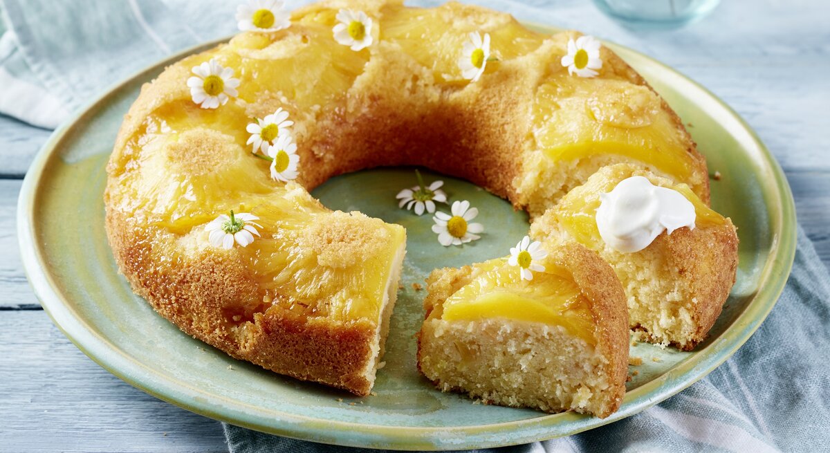 Auf einem Porzellanteller liegt ein Ananas-Kokos-Kuchen in Kranzform. Er ist mit weißen essbaren Blüten dekoriert. 