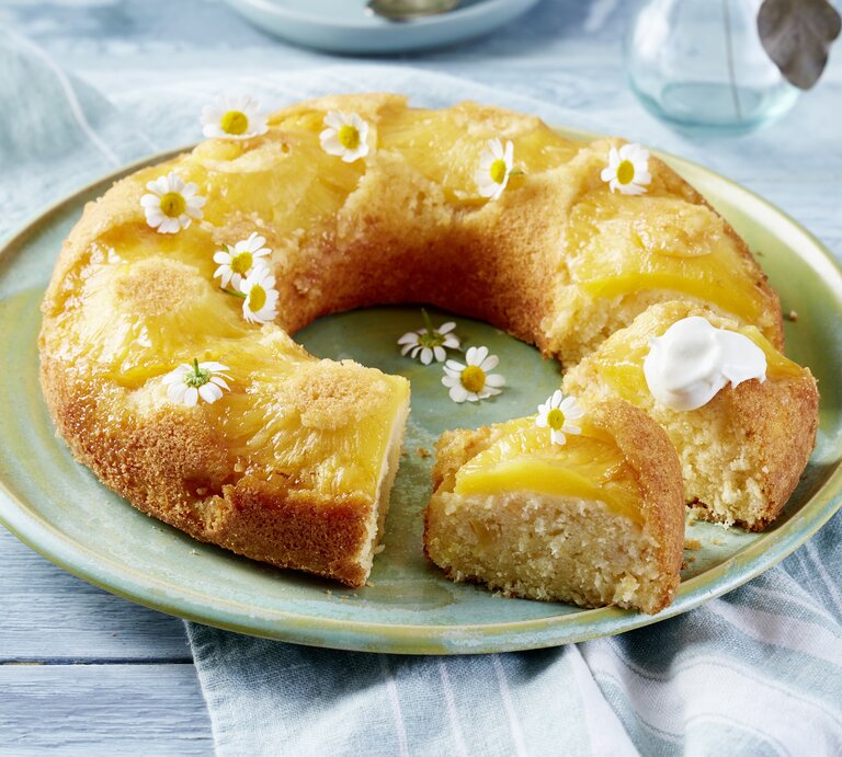 Auf einem Porzellanteller liegt ein Ananas-Kokos-Kuchen in Kranzform. Er ist mit weißen essbaren Blüten dekoriert.  | © EAT CLUB/Isabell Triemer