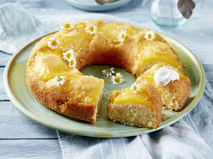Auf einem Porzellanteller liegt ein Ananas-Kokos-Kuchen in Kranzform. Er ist mit weißen essbaren Blüten dekoriert.  | © EAT CLUB/Isabell Triemer
