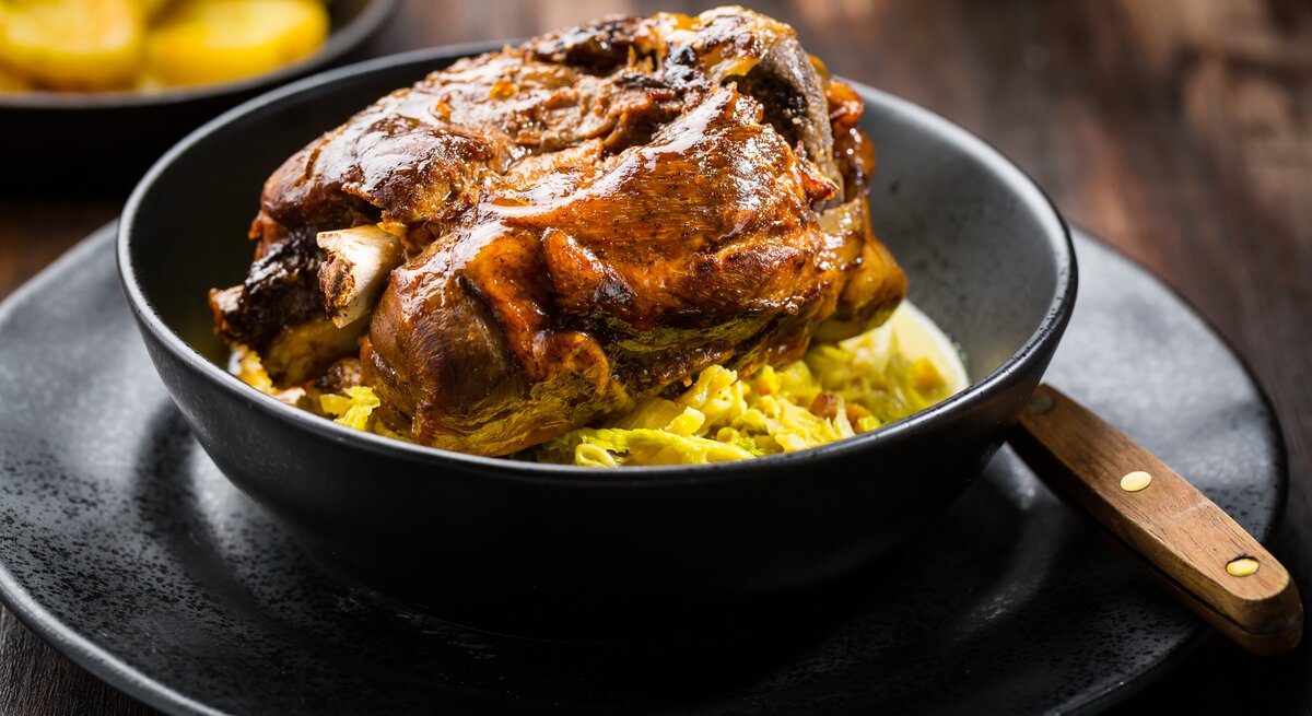 Frontal: Auf einem dunklen Untergrund stehen ein schwarzer Teller mit einer schwarzen Schüssel. Darin ist eine Bayerische Schweinshaxe auf sauerkarut.