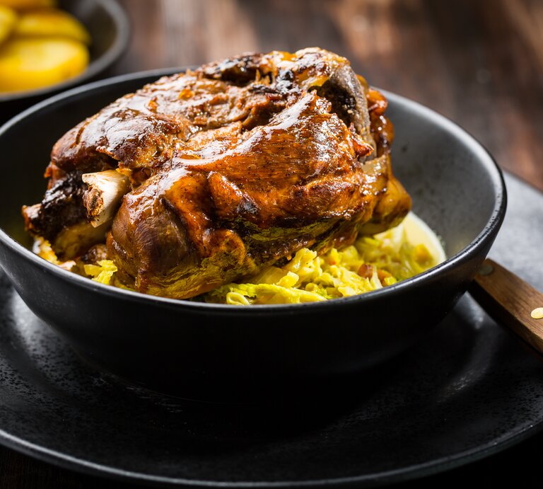 Frontal: Auf einem dunklen Untergrund stehen ein schwarzer Teller mit einer schwarzen Schüssel. Darin ist eine Bayerische Schweinshaxe auf sauerkarut. | © Adobe Stock / Brebca