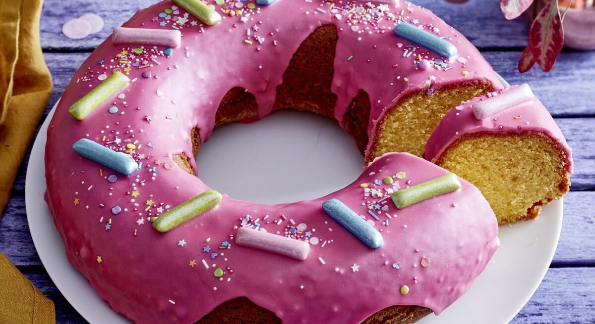 Auf einem Teller liegt ein  Donut-Kuchen in Kranzform. Er ist mit Glasur überzogen und mit bunten Streuseln und Süßigkeiten dekoriert.