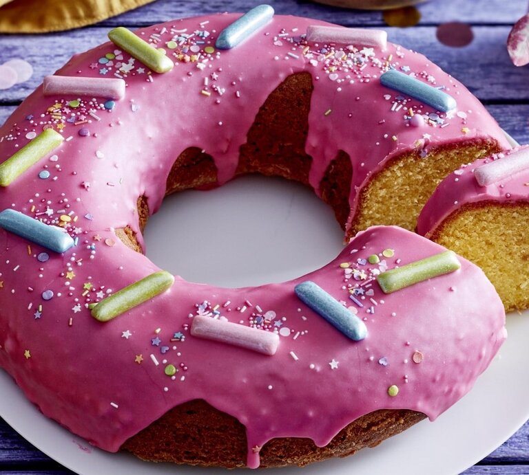 Auf einem Teller liegt ein  Donut-Kuchen in Kranzform. Er ist mit Glasur überzogen und mit bunten Streuseln und Süßigkeiten dekoriert. | ©  EAT CLUB / Isabell Triemer