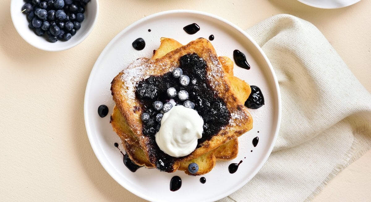 Draufsicht: Ein weißer Teller mit French Toast mit Blaubeeren und Vanille-Schmand liegt auf einem hellen Untergrund. Daneben liegen ein helles, gobes Tuch sowie einen Schale mit frischen Heidelbeeren.