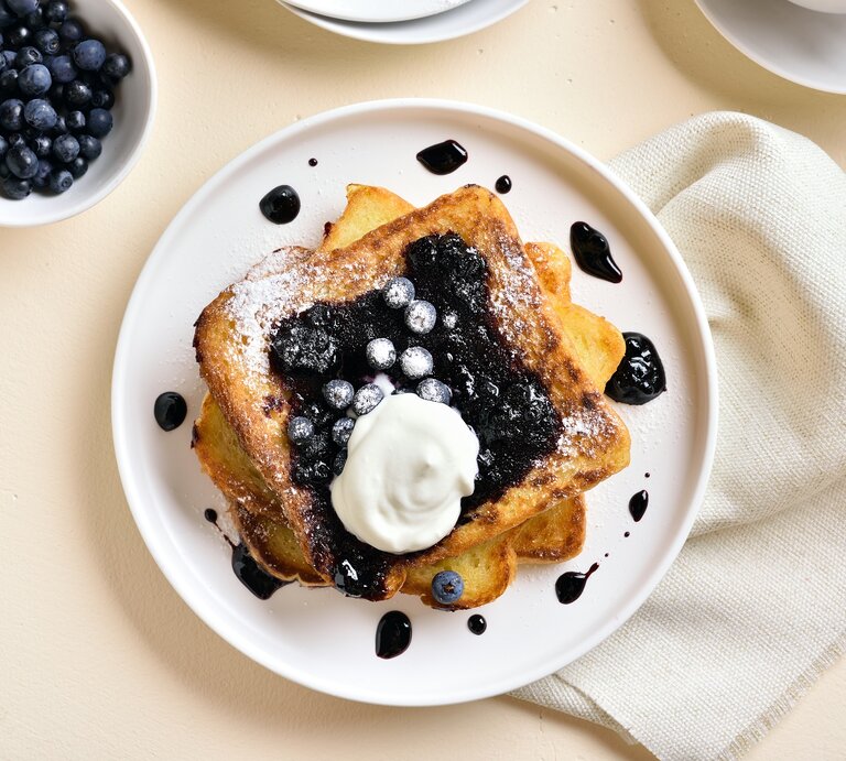 Draufsicht: Ein weißer Teller mit French Toast mit Blaubeeren und Vanille-Schmand liegt auf einem hellen Untergrund. Daneben liegen ein helles, gobes Tuch sowie einen Schale mit frischen Heidelbeeren. | © Shutterstock / Tatiana Volgutova