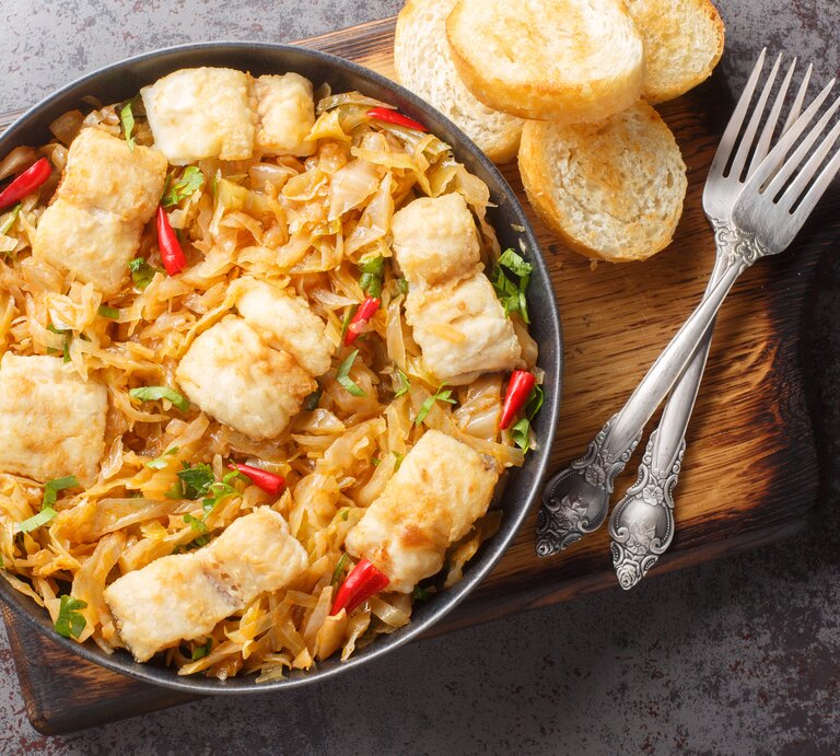 Sauerkrautauflauf mit Seelachs in schwarzer Schale auf Holzbrett. Daneben liegen Brotscheiben und Besteck. | © AdobeStock/FomaA