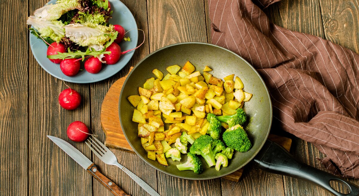 Draufsicht: Untergrund aus Holz, eine Pfanne  Bratkartoffeln mit Brokkoli sowie ein Salat mit Radieschen und Lollo Rosso-Salat. 