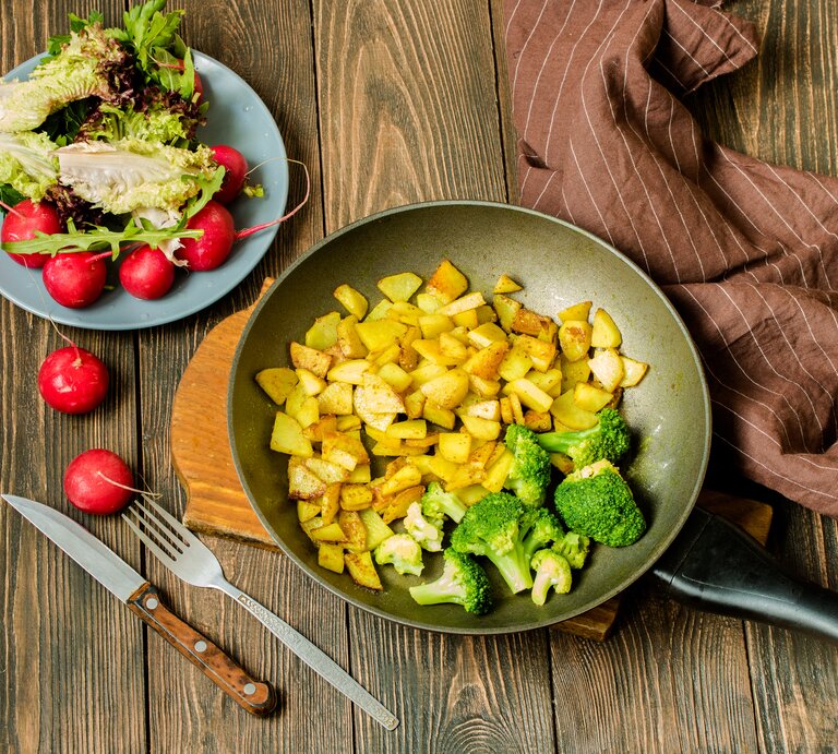 Draufsicht: Untergrund aus Holz, eine Pfanne  Bratkartoffeln mit Brokkoli sowie ein Salat mit Radieschen und Lollo Rosso-Salat.  | © Shutterstock / Sarkar Nataliia