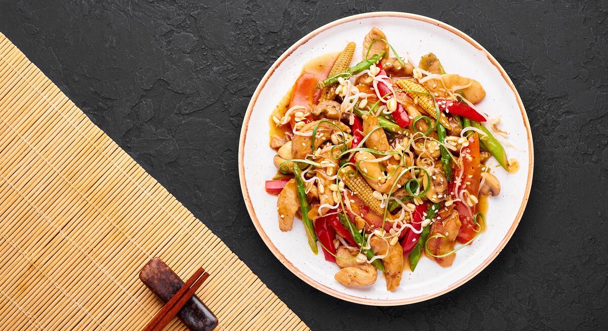 Ein weißer Teller mit dem Chop Suey auf dunklem Untergrund mit Stäbchen und Bambusmatte unten rechts von oben fotografiert.  