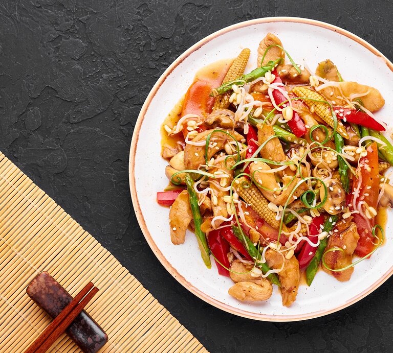Ein weißer Teller mit dem Chop Suey auf dunklem Untergrund mit Stäbchen und Bambusmatte unten rechts von oben fotografiert.   | © Shutterstock/ Kravtzov 
