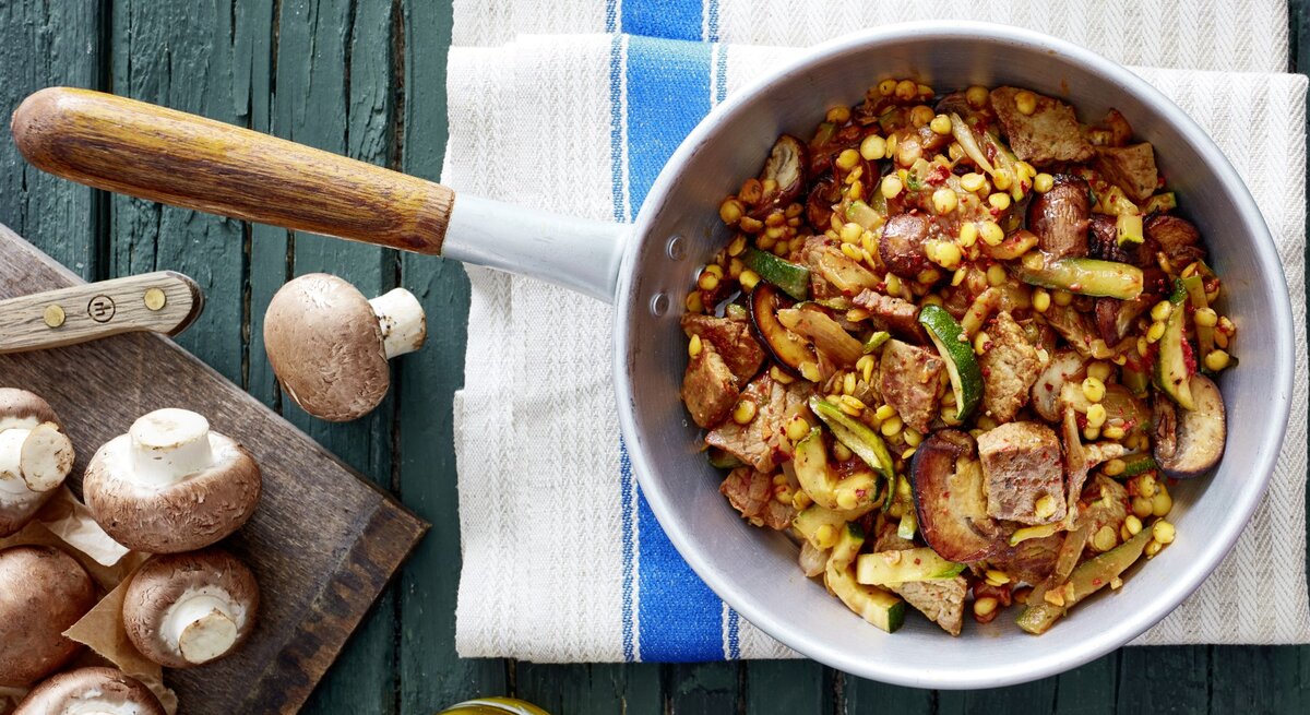 Schnitzel-Gemüse-Pfanne mit Linsen in Pfanne auf Küchentuch, daneben Flasche Öl und Champignons auf Brettchen, Untergrund dunkelgrünes Holz. 