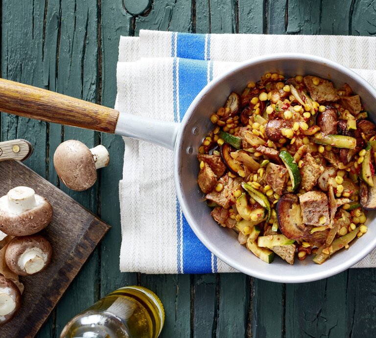 Schnitzel-Gemüse-Pfanne mit Linsen in Pfanne auf Küchentuch, daneben Flasche Öl und Champignons auf Brettchen, Untergrund dunkelgrünes Holz.  | © EAT CLUB / Isabell Triemer 