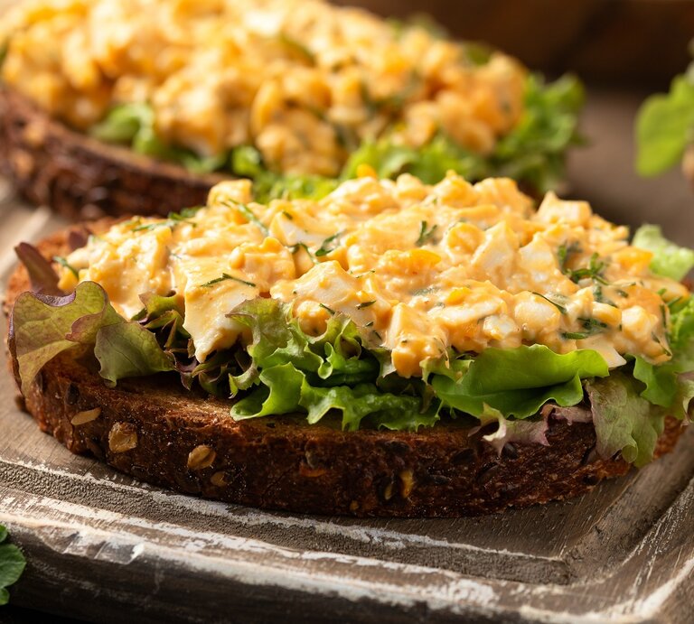 Zwei Brote mit dem Eiersalat auf Salatblättern. Alles auf einem dunkelbraunen Schneidebrett und rustikaler Tischplatte seitlich fotografiert. | © Shutterstock/ DronG 