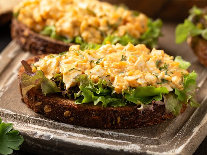 Zwei Brote mit dem Eiersalat auf Salatblättern. Alles auf einem dunkelbraunen Schneidebrett und rustikaler Tischplatte seitlich fotografiert. | © Shutterstock/ DronG 