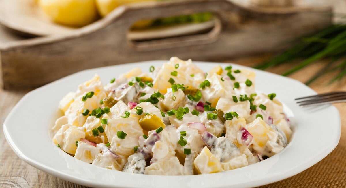 Ein Teller Kartoffelsalat mit Sour Cream, Joghurt, roten und Frühlingszwiebeln, im Hintergrund unscharf ein Tablett aus Holz mit einem Teller Kartoffeln. | © Adobe Stock/ gkrphoto