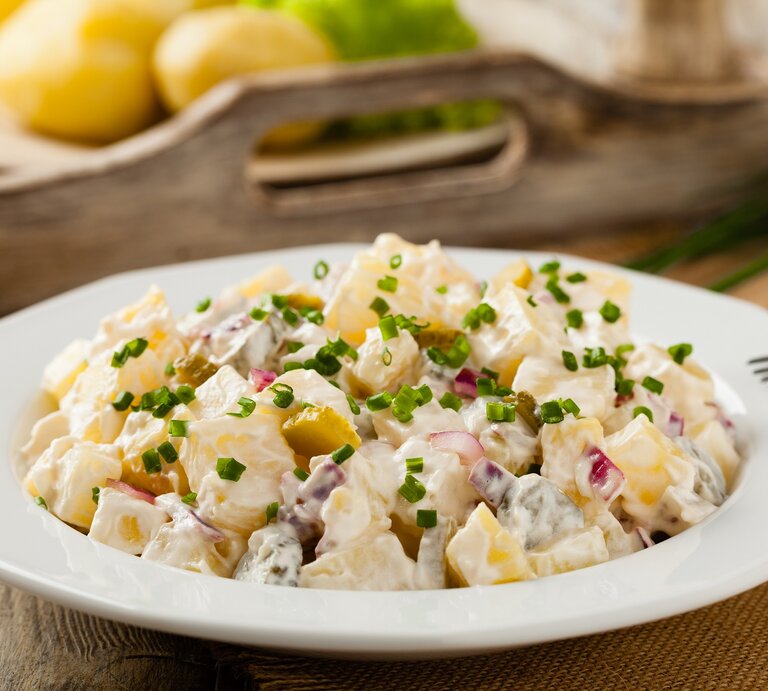 Ein Teller Kartoffelsalat mit Sour Cream, Joghurt, roten und Frühlingszwiebeln, im Hintergrund unscharf ein Tablett aus Holz mit einem Teller Kartoffeln. | © Adobe Stock/ gkrphoto