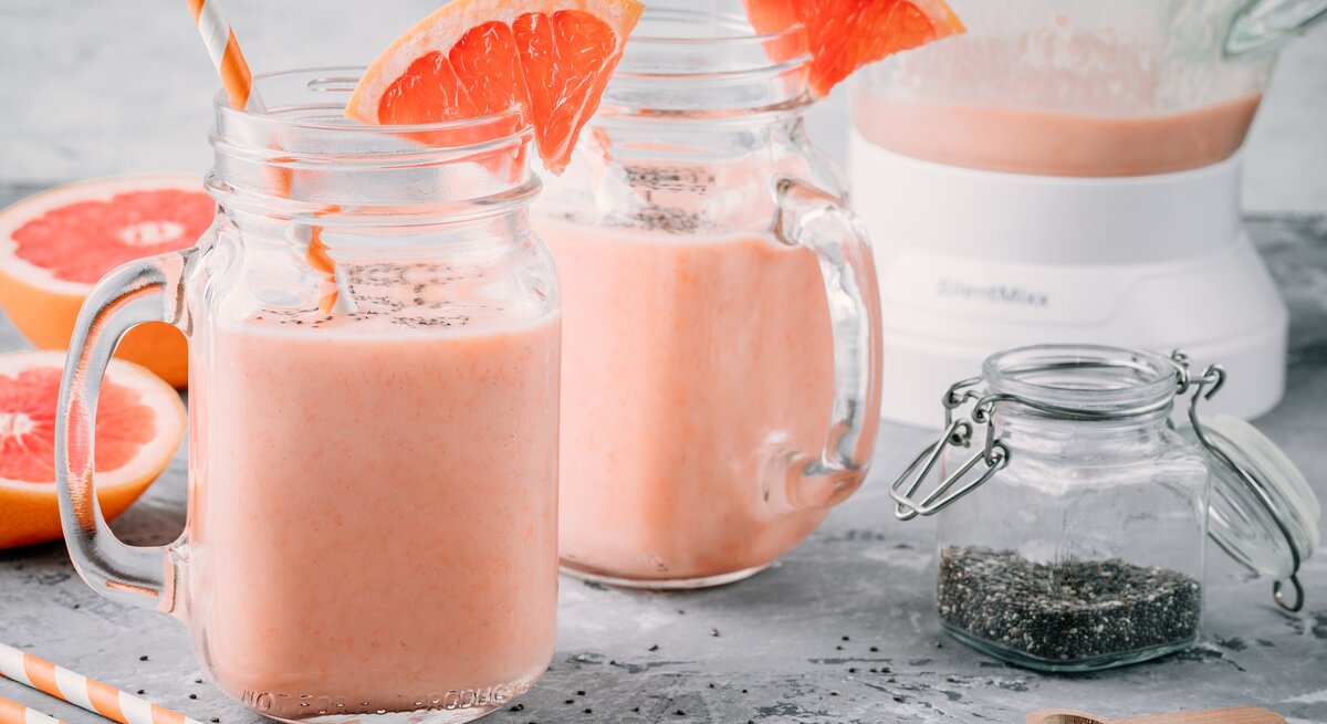 Frontalsicht: Auf grauem Untergrund stehen zwei Grapefruit-Chia-Smoothies, ein Glas mit Chia-Samen und ein Mixer. Daneben liegen zwei Hälften einer Grapefruit.
