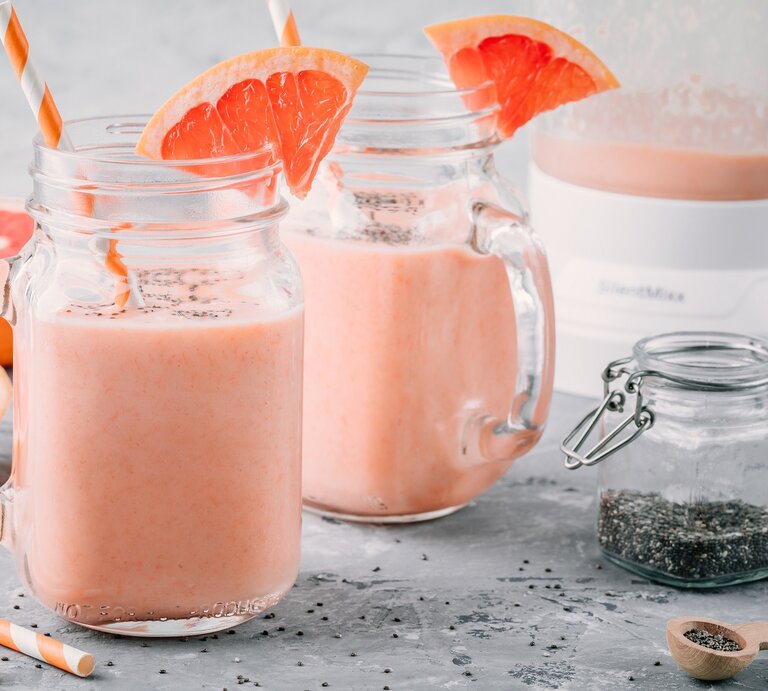 Frontalsicht: Auf grauem Untergrund stehen zwei Grapefruit-Chia-Smoothies, ein Glas mit Chia-Samen und ein Mixer. Daneben liegen zwei Hälften einer Grapefruit. | © Shutterstock / nblx