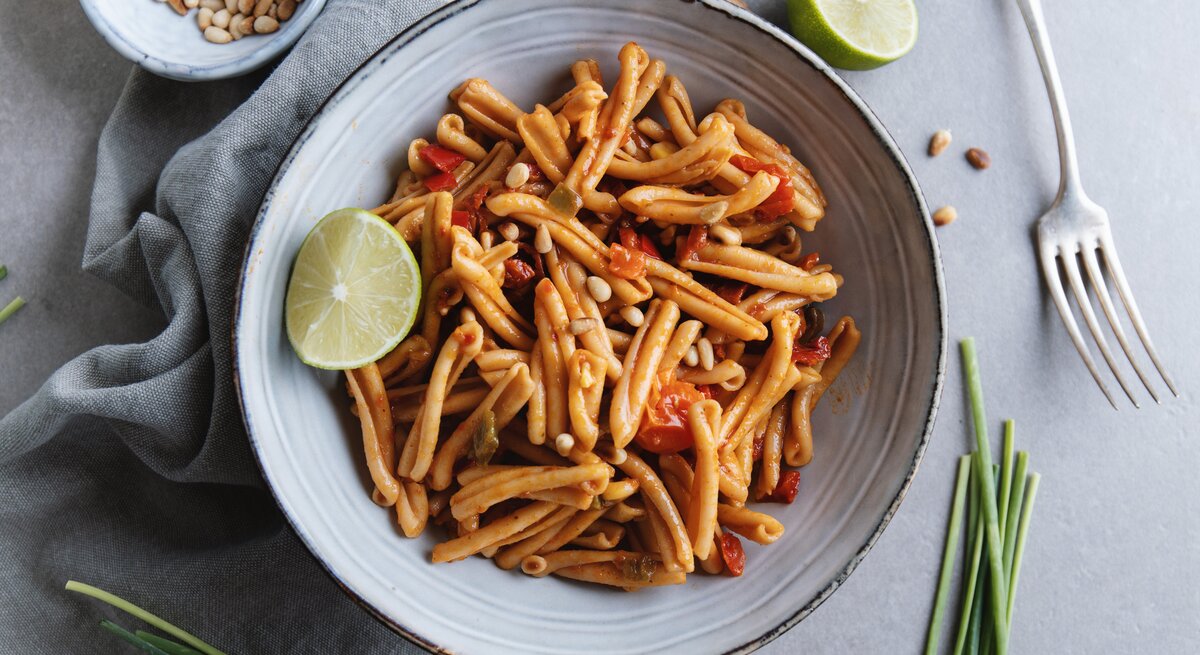 Tomaten-Pasta mit Pinienkernen dekoriert mit Limettenspalte auf weißem Teller. Daneben liegen Schnittlauch, Pinienkerne und eine Gabel. 