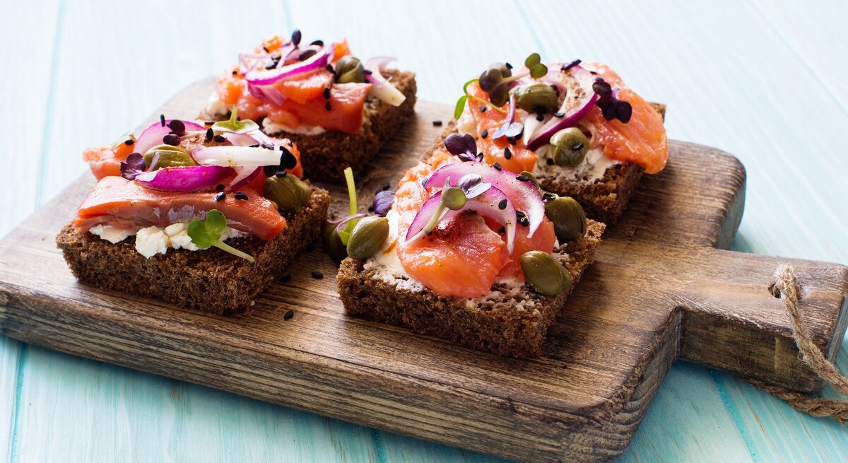 Vier Brote mit Lachs und Hüttenkäse auf einem dunklen Holzbrett und hellblauem Holzuntergrund von schräg oben fotografiert.