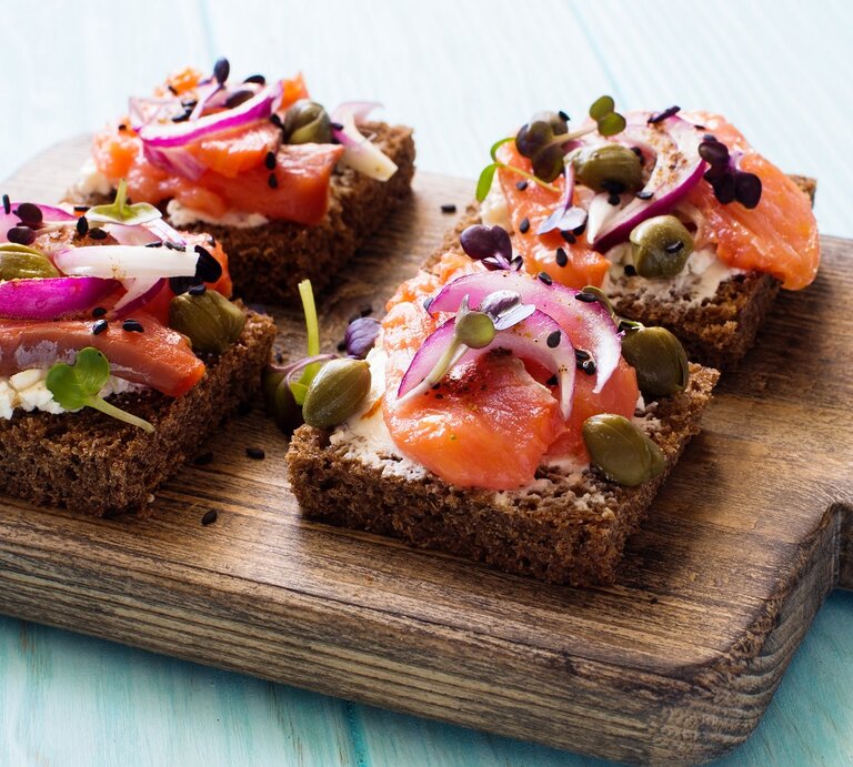 Vier Brote mit Lachs und Hüttenkäse auf einem dunklen Holzbrett und hellblauem Holzuntergrund von schräg oben fotografiert. | © Shutterstock/ annata78