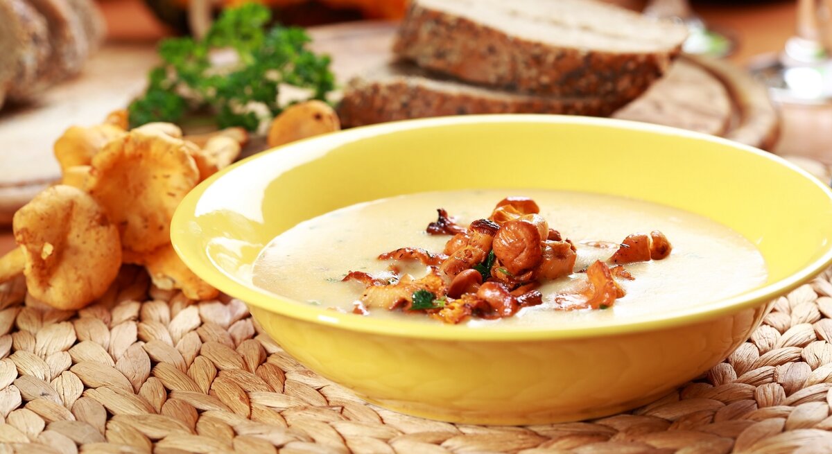 Ein gelber Suppenteller mit der Pfifferlingsuppe auf einer Bastmatte mit orange-gelbem Hintergrund. Außerdem Pfifferling, Brotscheiben und Petersilie im Hintergrund unscharf zu erkennen.