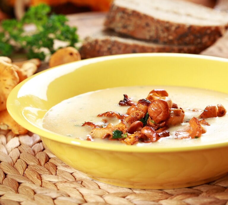 Ein gelber Suppenteller mit der Pfifferlingsuppe auf einer Bastmatte mit orange-gelbem Hintergrund. Außerdem Pfifferling, Brotscheiben und Petersilie im Hintergrund unscharf zu erkennen. | © Shutterstock/ Ingrid Balabanova 