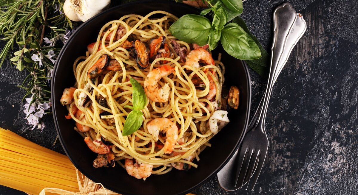 Ein schwarzer Teller auf grauem Tisch mit der Pasta. Daneben Kräuter, rohe Spaghetti und Besteck.