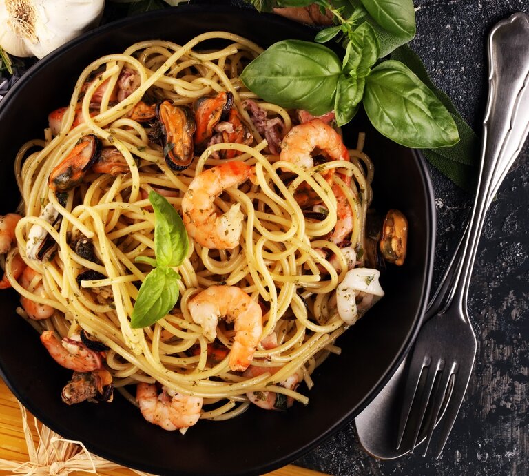 Ein schwarzer Teller auf grauem Tisch mit der Pasta. Daneben Kräuter, rohe Spaghetti und Besteck. | © Shutterstock/ beats1 