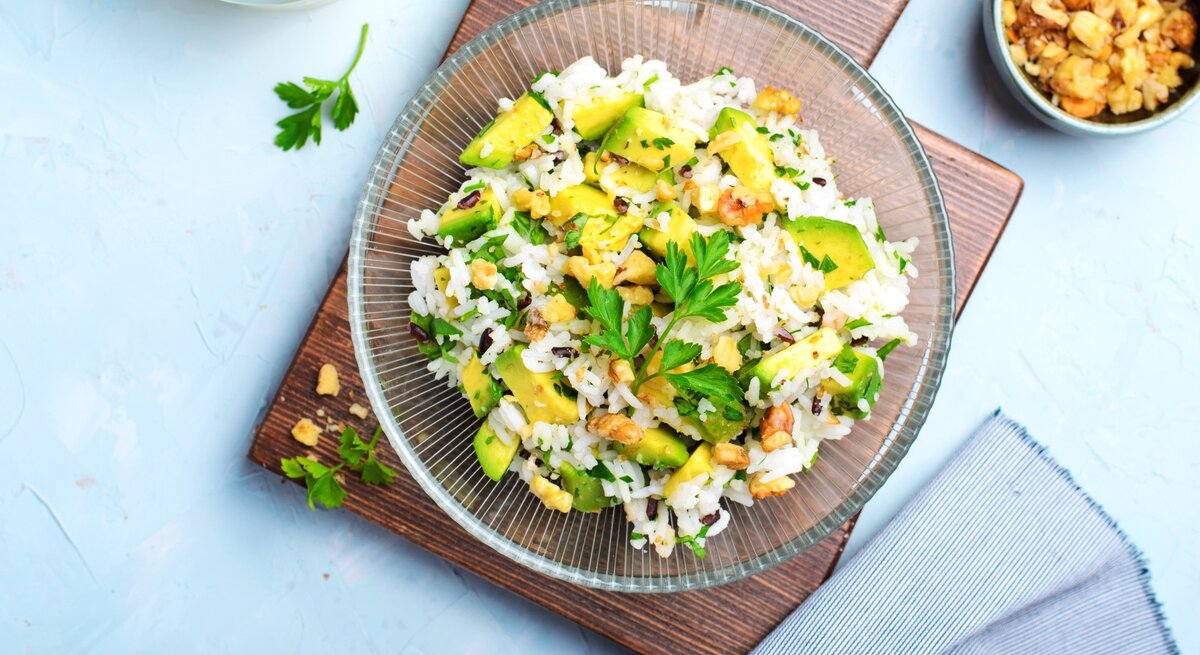 Draufsicht: Blauer Untergrund, ein quadratischtes Holzbrettchen, auf dem ein Teller aus Glas mit einem Avocado-Reissalat mit Mandeln liegt. Daneben liegt eine Schale mit ein paar gerösteten Mandeln.