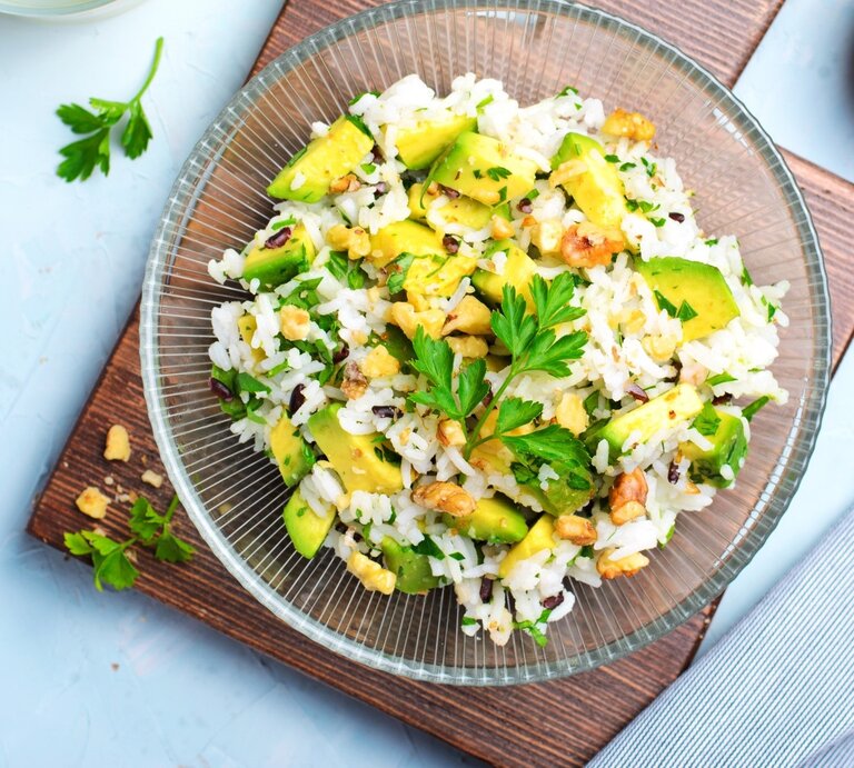 Draufsicht: Blauer Untergrund, ein quadratischtes Holzbrettchen, auf dem ein Teller aus Glas mit einem Avocado-Reissalat mit Mandeln liegt. Daneben liegt eine Schale mit ein paar gerösteten Mandeln. | © Getty Images / 	Iulia Cozlenco