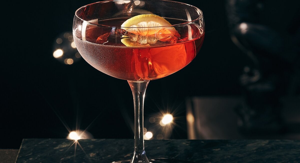 Frontal: Auf einer dunklen Tischplatte steht ein Glas Gin Dubonnet mit EIswürfeln und eine Zitronenschnitzer.