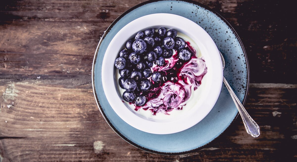 Draufsicht: Eine Schale Kokos-Granola mit Skyr und Blaubeeren auf einem blauen Teller auf dunklem Untergund aus Holz.