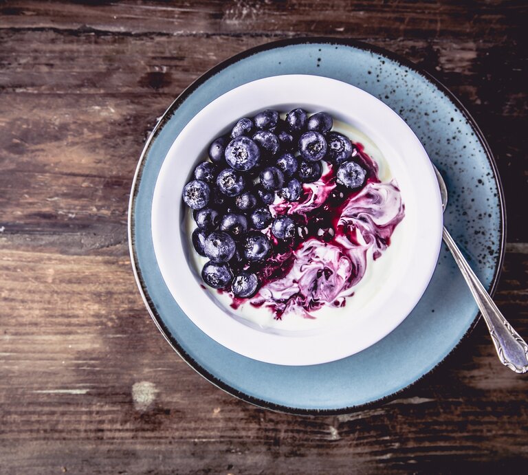 Draufsicht: Eine Schale Kokos-Granola mit Skyr und Blaubeeren auf einem blauen Teller auf dunklem Untergund aus Holz. | © Shutterstock /Fischer Food Design
