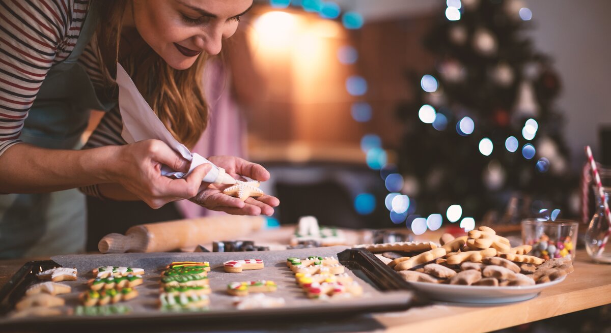 Frau verziert verschiedene ausgestochene Weihnachtsplätzchen, die auf einem Backblech liegen. 