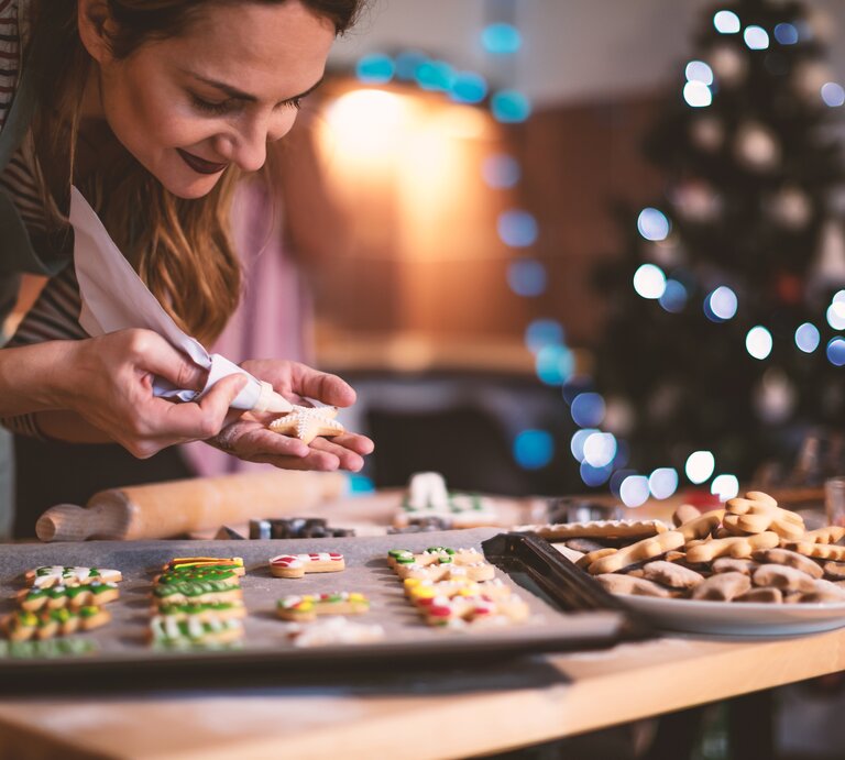 Frau verziert verschiedene ausgestochene Weihnachtsplätzchen, die auf einem Backblech liegen.  | © iStock.com/MilosStankovic