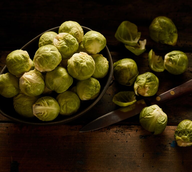 Rosenkohl  in dunkler Schale auf hölzernem Tisch, daneben ein Messer sowie einige angeschnittene Kohlköpfchen. | © AdobeStock/exclusive-design